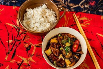 2 bowl, roasted beef with vegetable in spicy soy-chili sauce, rice and chopstick on red placemat, asian meal.