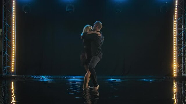 Beautiful couple dancing latin dance. Professional dancers in black clothes dance salsa on the surface of the water. Backlit dark studio. Slow motion.