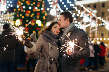 Couple in love having fun with sparklers on christmas decoration lights street. Romantic date for Valentine's day Outdoors. Young man and Woman laughing. Winter wonderland city  scene. New Year party