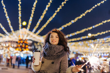 Girl having fun with sparklers on christmas decoration lights street. Young happy smilin Woman wearing stylish knitted scarf coat Outdoors. Model laughing. Winter wonderland city  scene New Year party