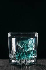 Gin in a glass on a black background with blue ice cubes on a wooden surface. Alcohol with ice..