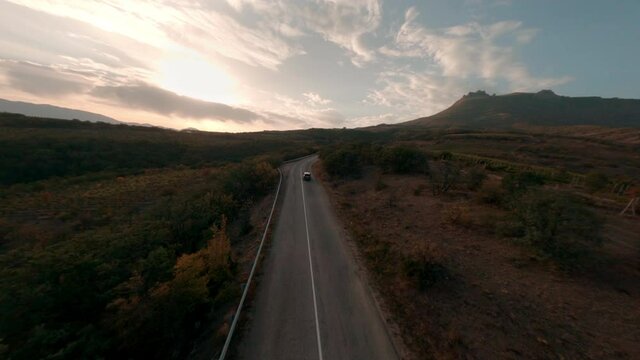 Black car cruising drives along empty asphalt road highway running across highland mountains valley under cloudy sunset sky in autumn sunset evening fpv sport drone first point aerial cinematic view.