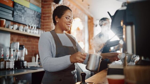Beautiful Latin American Female Barista with Short Hair and Glasses is Making a Cup of Tasty Cappuccino in Coffee Shop Bar. Male Cashier Works at a Cozy Loft-Style Cafe Counter in the Background.