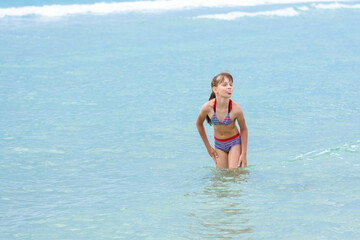 A girl stands knee-deep in sea water, does not want to go out to bask and sticks out her tongue