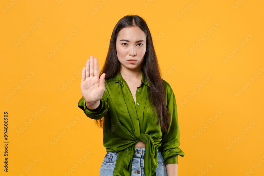 Wall mural Serious dissatisfied young brunette asian woman 20s wearing casual basic green shirt standing showing stop gesture with palm looking camera isolated on bright yellow colour background studio portrait. - Wall murals