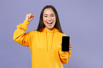 Joyful young brunette asian woman 20s in basic yellow hoodie standing hold mobile cell phone with blank empty screen doing winner gesture isolated on pastel violet colour background, studio portrait.