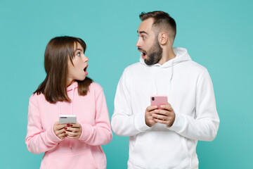 Shocked young couple two friends man woman 20s wearing white pink casual hoodie using mobile cell phone typing sms message looking at each other isolated on blue turquoise background studio portrait.