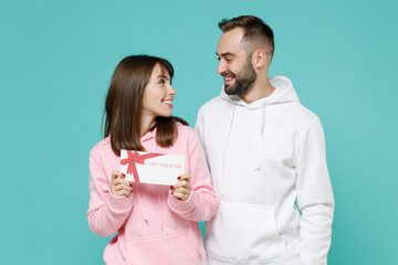 Smiling funny young couple two friends man woman 20s wearing white pink casual hoodie hold gift certificate looking at each other isolated on blue turquoise colour wall background studio portrait.