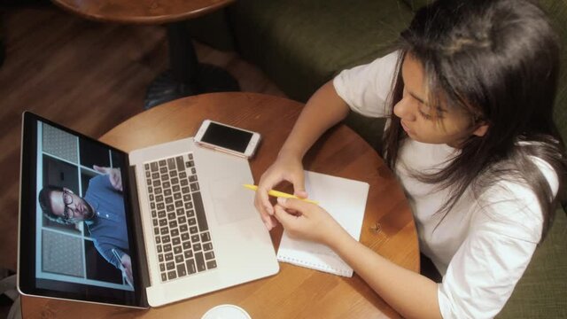 Young girl student watching lesson online and studying from home. Mixed race teenager taking notes while looking at computer screen. Girl student studying at home.