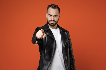 Strict irritated dissatisfied young bearded man wearing basic white t-shirt black leather jacket standing pointing index finger on camera isolated on bright orange colour background studio portrait.