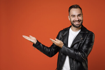 Smiling cheerful young bearded man wearing basic white t-shirt black leather jacket standing pointing hands aside on mock up copy space isolated on bright orange colour background studio portrait.