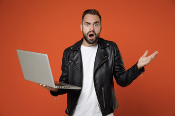 Shocked worried young bearded man wearing basic white t-shirt black leather jacket standing working on laptop pc computer spreading hands isolated on bright orange colour background studio portrait.