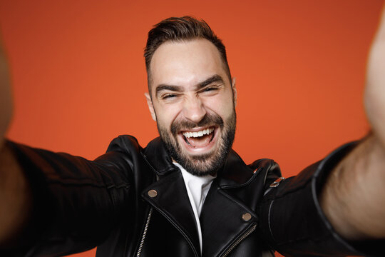 Close up of laughing young bearded man 20s wearing basic white t-shirt black leather jacket standing doing selfie shot on mobile phone looking camera isolated on orange background studio portrait.