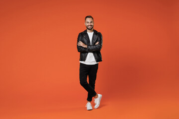 Fototapeta na wymiar Full length of smiling attractive young bearded man 20s wearing basic white t-shirt black leather jacket standing holding hands crossed looking camera isolated on orange background studio portrait.
