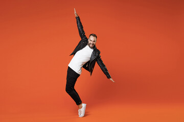 Fototapeta na wymiar Full length side view of laughing young bearded man in basic white t-shirt black leather jacket standing on toes dancing rising spreading hands isolated on orange colour background studio portrait.