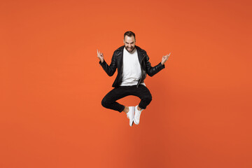 Fototapeta na wymiar Full length of cheerful young bearded man 20s in basic white t-shirt black leather jacket jumping depicting heavy metal rock sign looking camera isolated on orange colour background studio portrait.