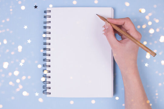 Christmas planning concept. A girl writing in a notebook with clean pages. Christmas decor, lights, design paper. Top view.