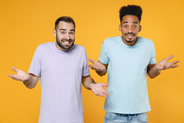 Confused puzzled young friends european african american men 20s wearing casual violet blue t-shirts spreading hands looking camera isolated on bright yellow colour wall background studio portrait.