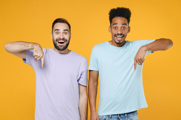 Surprised excited young friends european african american men 20s wearing casual violet blue t-shirts standing pointing index fingers down isolated on bright yellow colour background studio portrait.