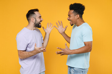 Side view of cheerful joyful young friends european african american men 20s in casual violet blue t-shirts spreading hands looking at each other isolated on yellow colour background studio portrait.