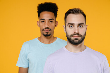 Fototapeta na wymiar Handsome serious young two friends european african american men 20s wearing violet blue casual t-shirts standing and looking camera isolated on bright yellow colour wall background studio portrait.