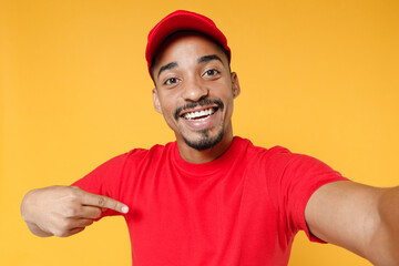 Fototapeta premium Close up fun delivery employee african man in red cap blank print tshirt uniform workwear work courier dealer service concept doing selfie shot on mobile phone isolated on yellow background studio