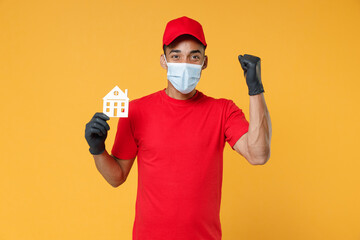 Fototapeta na wymiar Delivery employee african man in red cap print t-shirt face mask gloves uniform work courier service to home on quarantine coronavirus covid-19 virus concept hold house isolated on yellow background.
