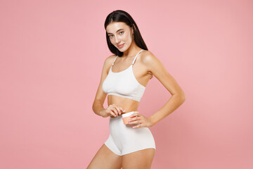 Fototapeta na wymiar Side view of smiling attractive beautiful young brunette woman 20s in white underwear posing hold container with moisturizer body cream isolated on pastel pink colour wall background, studio portrait.