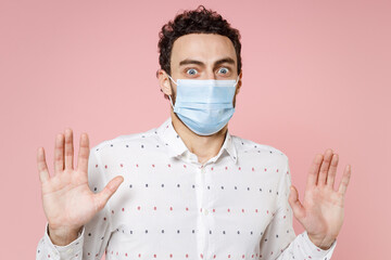 Shocked young bearded man 20s in basic white shirt sterile face mask safe from coronavirus virus covid-19 during quarantine rising hands up showing palms isolated on pink background studio portrait.
