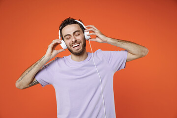 Fototapeta na wymiar Cheerful laughing funny young bearded man 20s wearing casual basic violet t-shirt listening music with headphones keeping eyes closed isolated on bright orange color wall background studio portrait.