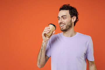 Cheerful smiling handsome young bearded man 20s wearing casual basic violet t-shirt standing hold paper cup of coffee or tea looking aside isolated on bright orange color background studio portrait.