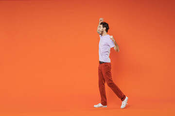 Fototapeta na wymiar Full length side view of excited joyful young bearded man 20s in casual violet t-shirt standing doing winner gesture clenching fists isolated on bright orange color wall background studio portrait.
