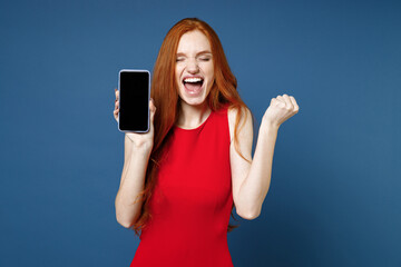 Happy young redhead woman 20s wearing bright red elegant evening dress standing hold mobile cell phone with blank empty screen doing winner gesture isolated on blue color background studio portrait.