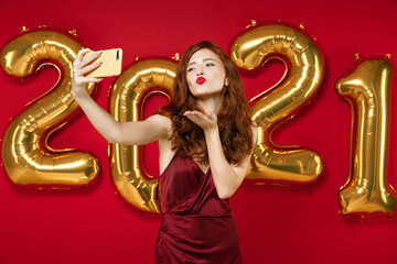 Cute young woman in elegant dress doing selfie shot on mobile phone blowing sending air kiss isolated on red background, golden numbers air balloons. Happy New Year 2021 celebration holiday concept.