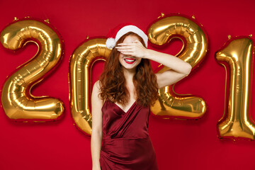 Cheerful young Santa woman in elegant dress Christmas hat covering eyes with hand hiding isolated on red background golden numbers air balloons. Happy New Year 2021 celebration holiday party concept.