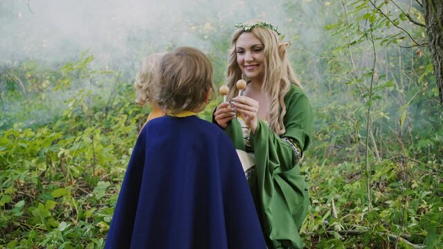 Fairy elf cosplay young woman treats little babies boys hobbits with candies in green forest. Halloween concept, fairytale characters, kids.
