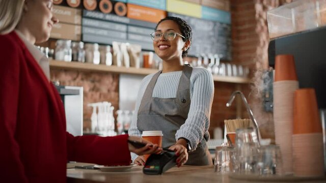 Joyful Multiethnic Diverse Woman Gives a Payment Terminal to Customer Using NFC Technology on Smartphone. Customer Uses Mobile to Pay for Take Away Latte and Pastry to a Barista in Coffee Shop.