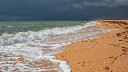 Endless yellow sandy beach with white foam waves under a dark stormy sky, panoramic