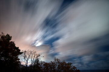 The movement of clouds in the night moonlit sky on a long exposure above the forest silhouette