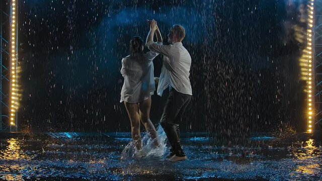 Wet man and a woman in white shirts are passionately dancing salsa, their bodies tightly pressed against each other. The couple dances on the surface of the water creating many splashes. Slow motion.