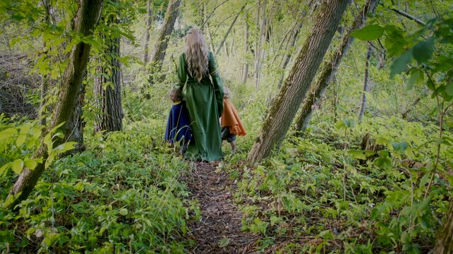 Two little toddler boys cosplay gnomes or hobbits with elf woman walking in green forest. Unrecognizable fairy tale characters. Halloween, kids concept. Amazing light. Slow motion. 