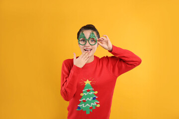 Young woman in Christmas sweater and party glasses on yellow background