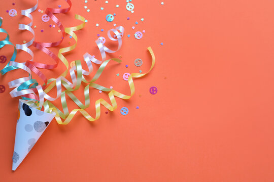 Colorful confetti and streamers with party cracker on orange background, top view. Space for text