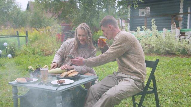 The husband drinks beer and shows his wife funny photos on a smartphone at the dacha during lunch