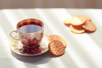 A cup of tea in a beautiful vintage tea cup and some biscuits on the table. Sun shines onto the table through curtains makin beautiful shadows.