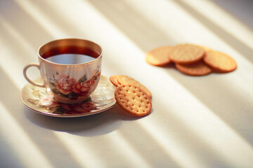 A cup of tea in a beautiful vintage tea cup and some biscuits on the table. Sun shines onto the table through curtains makin beautiful shadows.