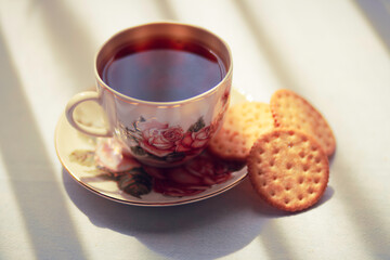 A cup of tea in a beautiful vintage tea cup and some biscuits on the table. Sun shines onto the table through curtains makin beautiful shadows.