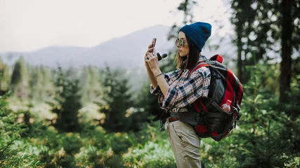 Tuinposter Stylish woman traveler in a hat stands on the top of the mountain. Hipster girl on the top of the mountain, resting and taking pictures on the phone. Atmospheric moment. Wanderlust and travel concept © Monako Art Studio