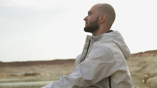 Side view of handsome bald bearded man chilling out with sandboard in desert or sand quarry