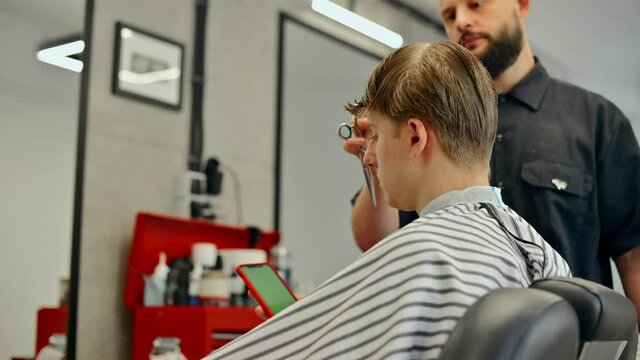 The client sits in the chair of a men's hairdresser, gets a barber's haircut and uses a smartphone. Barber cuts a business young man with a smartphone in hand